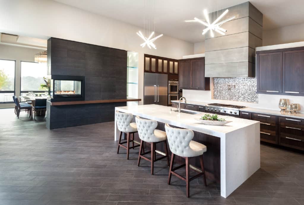 A bright, modern kitchen with a white island, three bar stools, dark cabinets, stainless steel appliances and starbursts lights