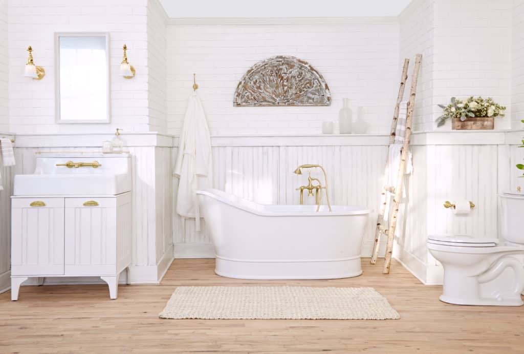 White bathroom with freestanding tub, wainscoting, gold fixtures, and wood floor