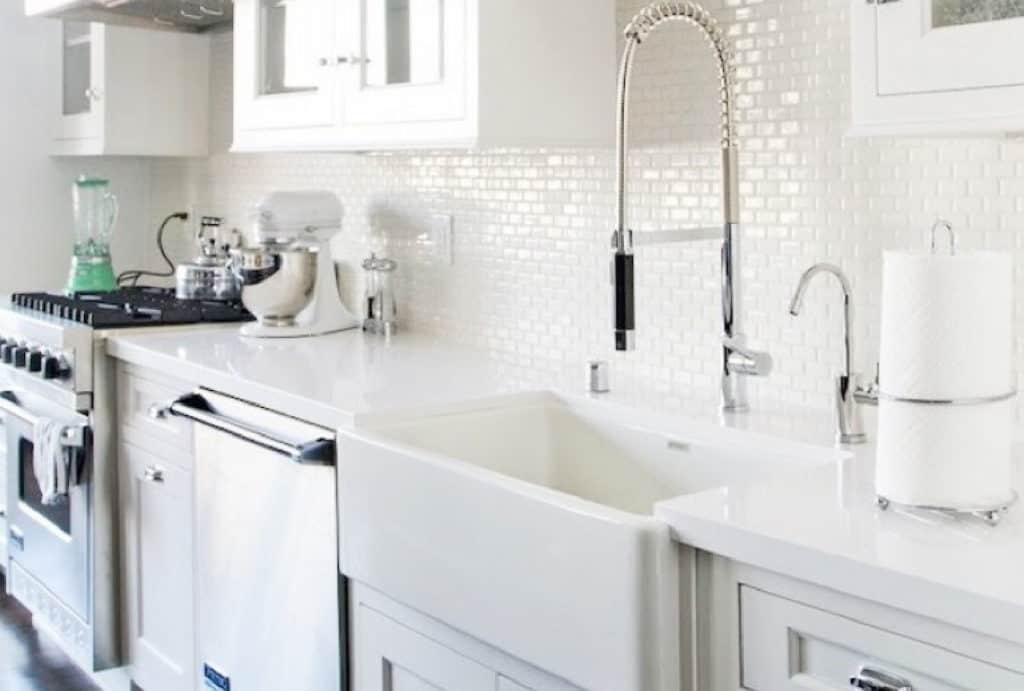 Bright white kitchen featuring a farmhouse sink with a modern faucet, white countertops, textured white backsplash, white cabinets, and stainless steel appliances