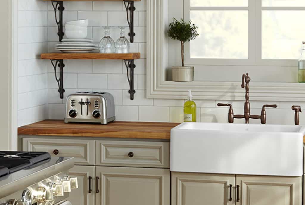 This charming kitchen from Coburn’s kitchen design services features open shelves, a wooden countertop with a toaster, a white farmhouse sink with a bronze faucet, and light gray cabinets against a white subway tile backsplash.
