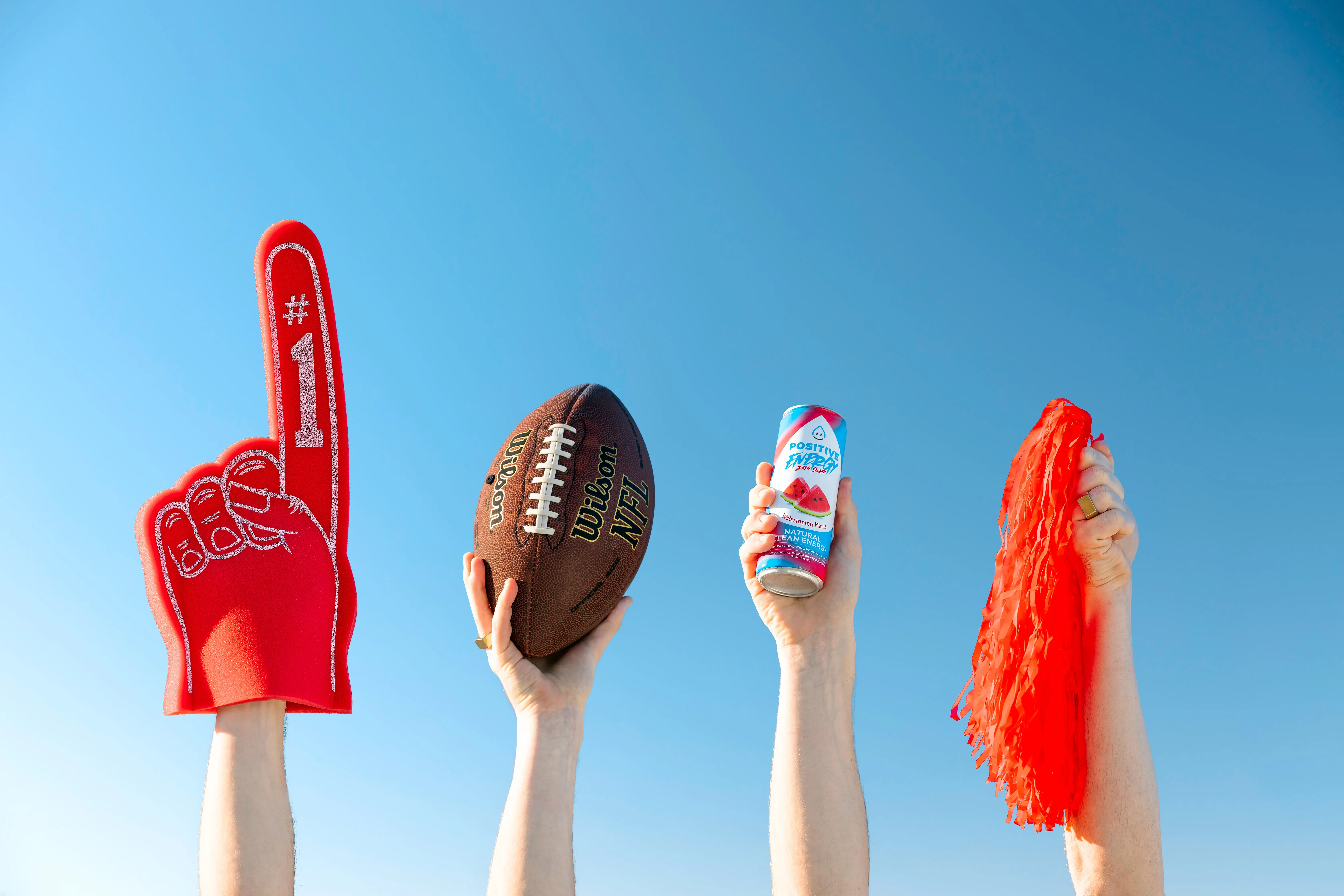 4 arms raised in the air holding a foam finger, a football, a drink can and a pom pom for a football party