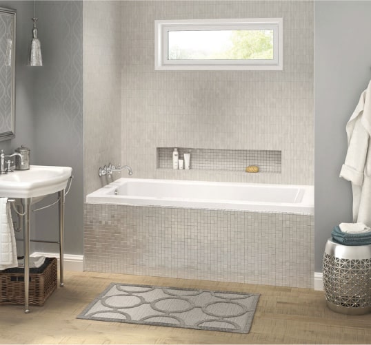 an alcove tub lined with grey tile in a bathroom accented with grey and light wood. Find more bathroom design ideas from Coburn's Kitchen & Bath Showroom.