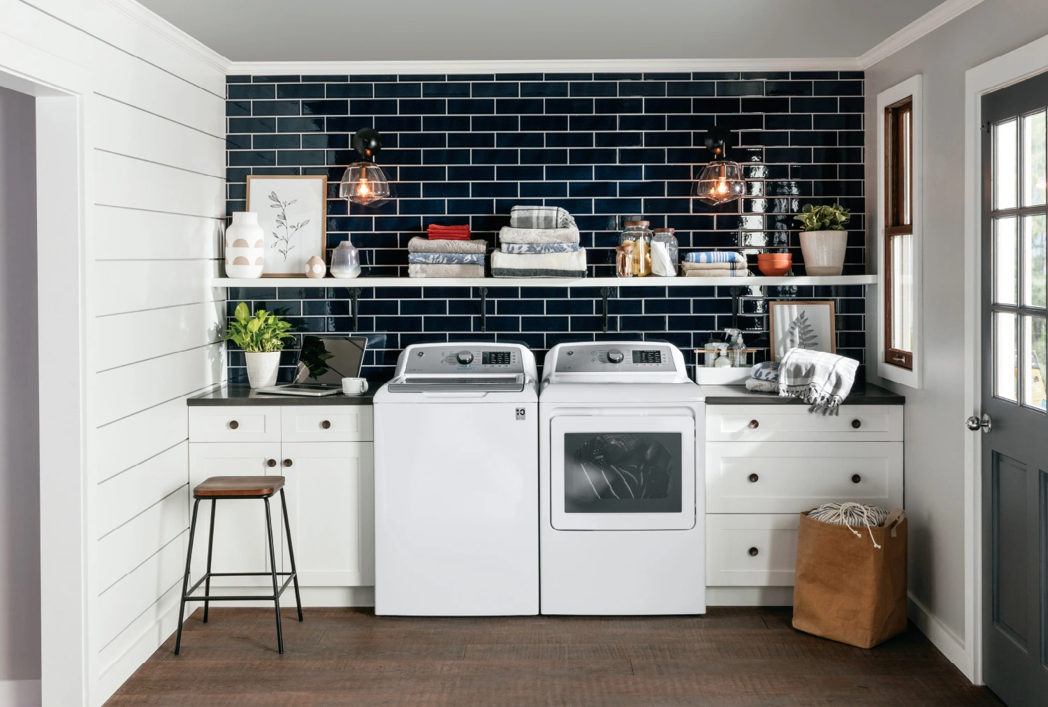 Luxury laundry room design ideas from Coburn’s Kitchen & Bath Showroom. Featuring a washer and dryer set, black tile backsplash, laundry room shelving, cabinets and counter space for additional laundry room storage options.